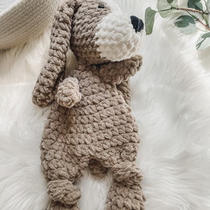 May include: A crocheted stuffed dog toy with brown and white fur. The dog is lying on a white fluffy surface.