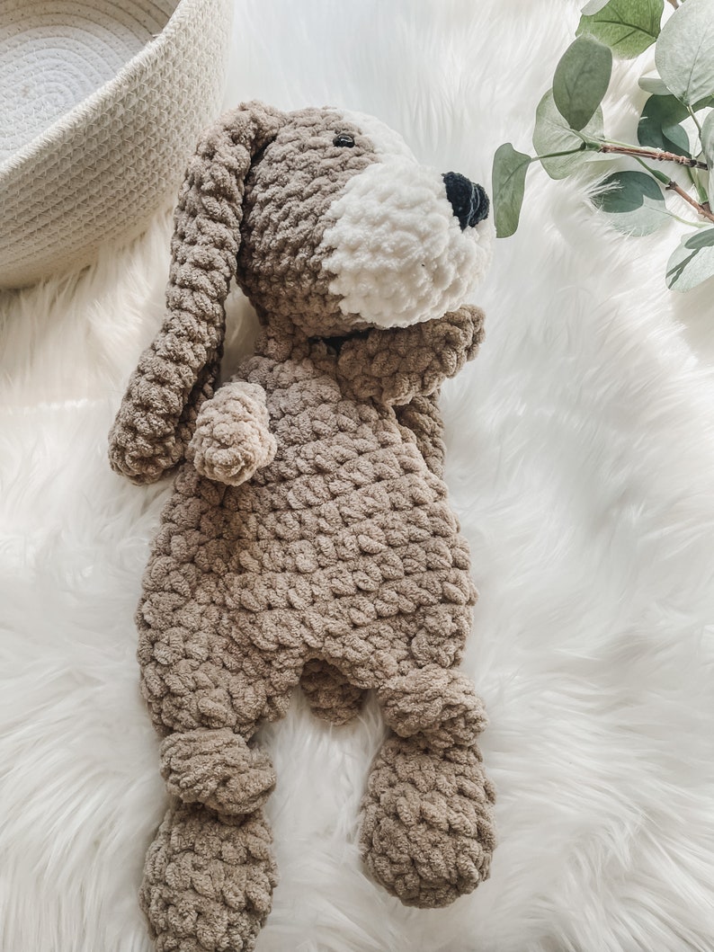 May include: A crocheted stuffed dog toy with brown and white fur. The dog is lying on a white fluffy surface.