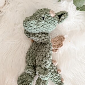 Peut inclure: Une peluche dragon en crochet vert faite &agrave; la main, avec des ailes et des accents beiges. Le dragon a une apparence textur&eacute;e et &eacute;paisse et est pos&eacute; sur une surface blanche et moelleuse. La t&ecirc;te du dragon pr&eacute;sente de petits yeux noirs et une tache rose.