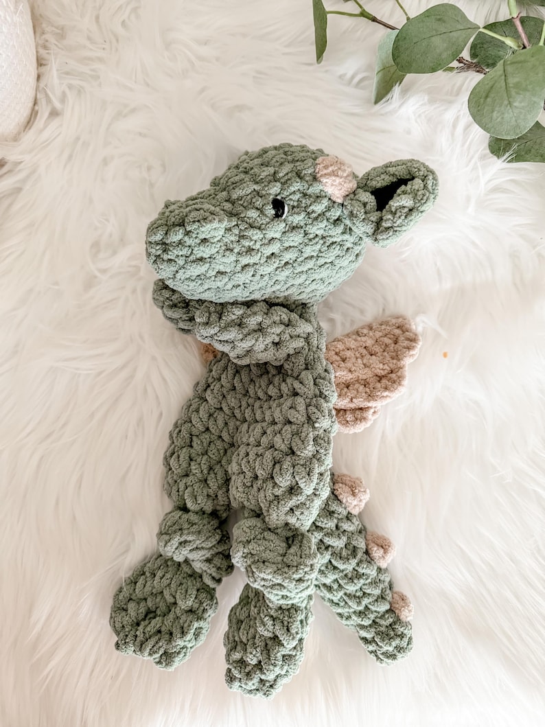 Peut inclure: Une peluche dragon en crochet vert faite &agrave; la main, avec des ailes et des accents beiges. Le dragon a une apparence textur&eacute;e et &eacute;paisse et est pos&eacute; sur une surface blanche et moelleuse. La t&ecirc;te du dragon pr&eacute;sente de petits yeux noirs et une tache rose.