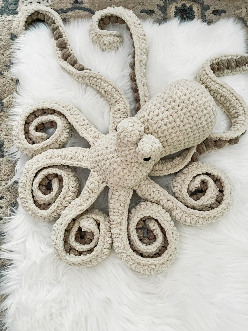 Pode incluir: Um brinquedo de pel&uacute;cia de polvo de croch&ecirc; com um corpo bege e tent&aacute;culos marrons. O polvo est&aacute; deitado em uma superf&iacute;cie branca e fofa.