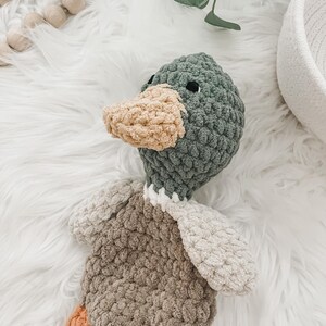 May include: A crocheted plush duck toy with a green head, brown body, and orange feet. The duck is lying on a white fluffy surface.