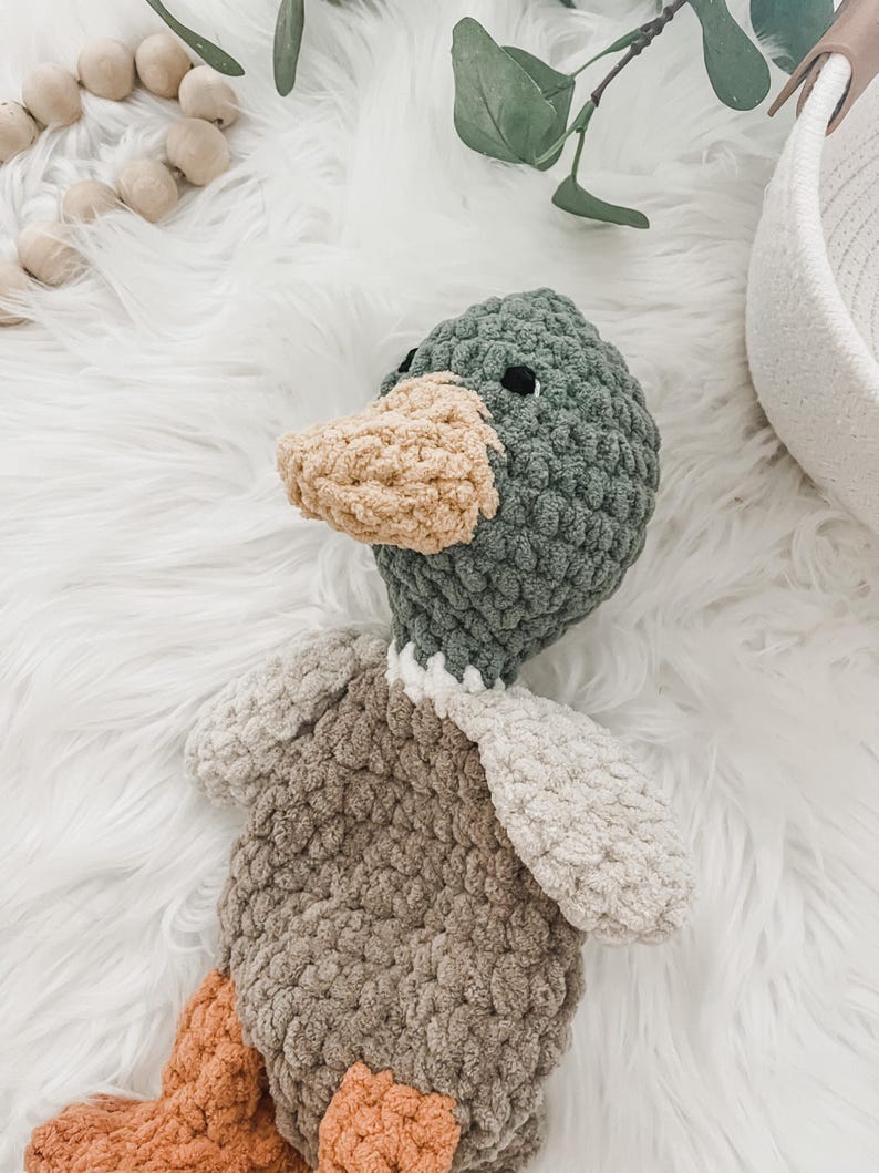 May include: A crocheted plush duck toy with a green head, brown body, and orange feet. The duck is lying on a white fluffy surface.