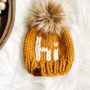 May include: A mustard yellow knitted beanie with a white "Hi" embroidered on the front. The beanie has a brown faux fur pom pom on top and a brown leather tag with the text "The Knotted Baby" on the side.
