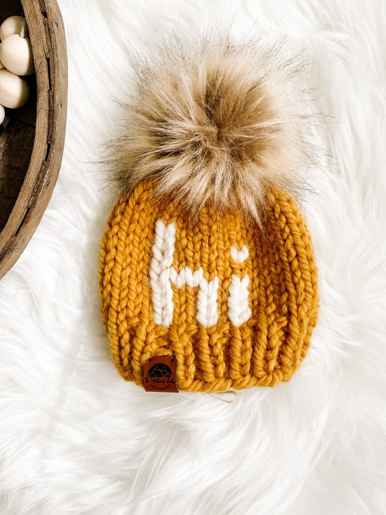 May include: A mustard yellow knitted beanie with a white "Hi" embroidered on the front. The beanie has a brown faux fur pom pom on top and a brown leather tag with the text "The Knotted Baby" on the side.