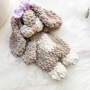 May include: A crocheted plush bunny toy with brown and white fur, wearing a purple bow. The bunny is lying on a white fluffy surface.