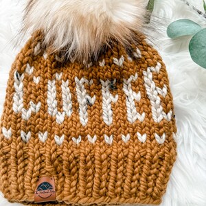 May include: A chunky knit beanie in a warm, golden-brown color, adorned with a cream-colored pattern and a fluffy, light brown pom-pom. The hat features a small leather tag with a mountain and tree logo. The beanie is a winter accessory.