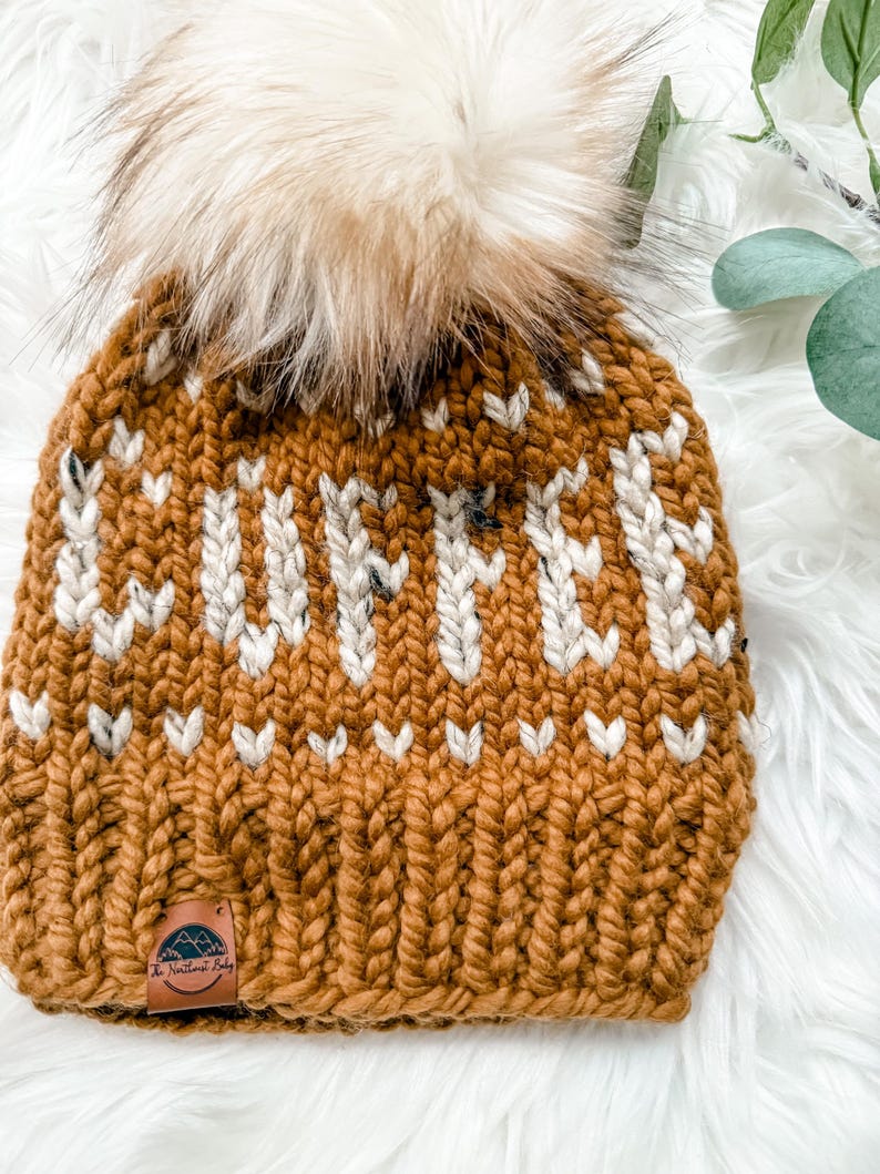 May include: A chunky knit beanie in a warm, golden-brown color, adorned with a cream-colored pattern and a fluffy, light brown pom-pom. The hat features a small leather tag with a mountain and tree logo. The beanie is a winter accessory.