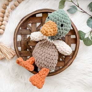 May include: A handmade crochet duck toy with a green head, orange beak, and brown body. The duck has white wings and orange feet. It is placed on a woven wooden tray with a white fluffy background. Decorative wooden beads and greenery are also present.