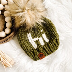 May include: A green knitted beanie with a white pom pom and a white "HI" embroidered on the front. The beanie has a brown leather tag with a logo on it.