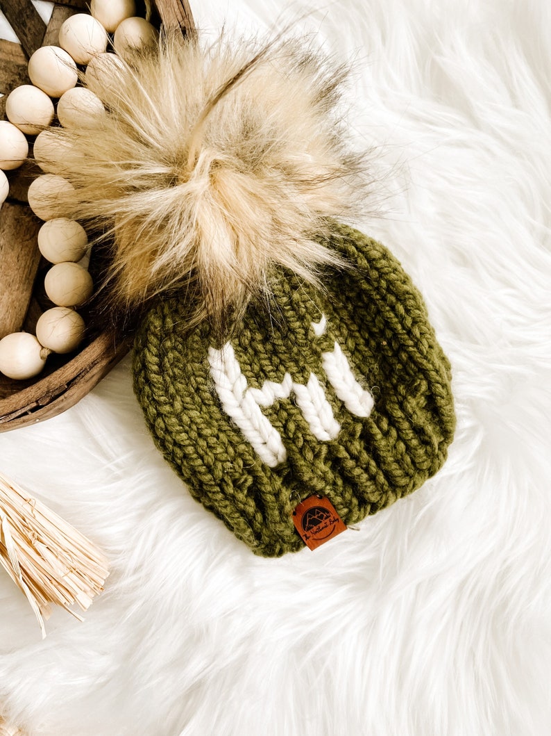 May include: A green knitted beanie with a white pom pom and a white "HI" embroidered on the front. The beanie has a brown leather tag with a logo on it.