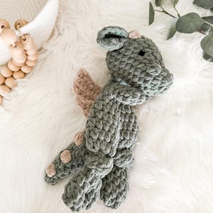 Peut inclure: Une peluche dragon faite &agrave; la main, crochet&eacute;e, dans des tons de gris et de beige. Le dragon pr&eacute;sente un corps textur&eacute;, de petites ailes et des taches d&eacute;coratives. Le jouet est con&ccedil;u avec une apparence douce et c&acirc;line.