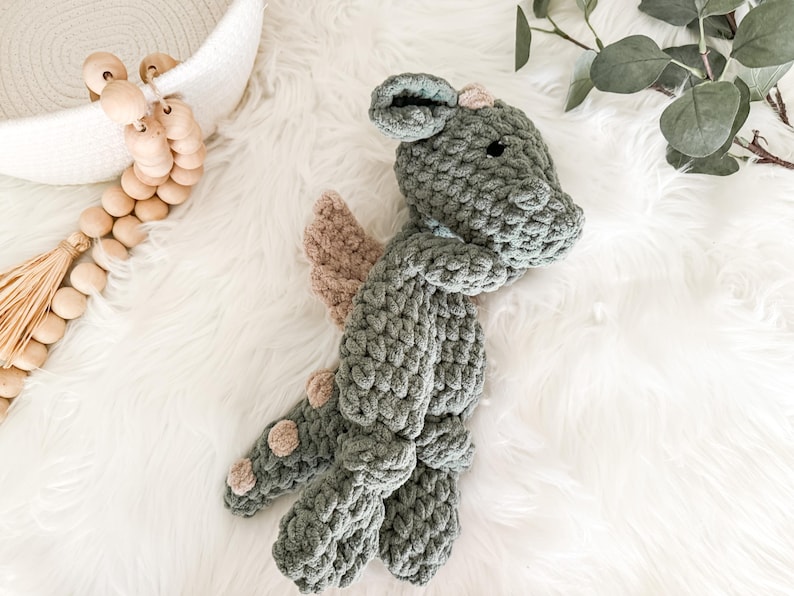 Peut inclure: Une peluche dragon faite &agrave; la main, crochet&eacute;e, dans des tons de gris et de beige. Le dragon pr&eacute;sente un corps textur&eacute;, de petites ailes et des taches d&eacute;coratives. Le jouet est con&ccedil;u avec une apparence douce et c&acirc;line.