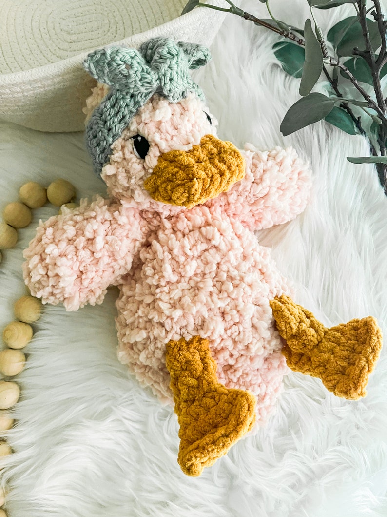 May include: A pink crocheted duck plush toy with a green bow on its head and yellow crocheted feet.