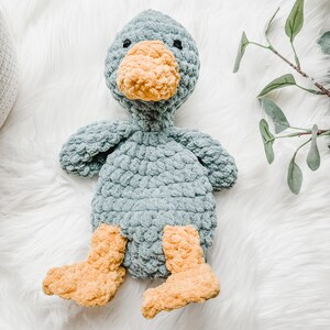 May include: A handmade crochet duck plush toy. The duck has a light blue body, a yellow-orange beak and feet, and small black eyes. The plush is sitting on a white, fluffy surface.