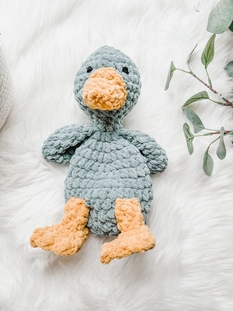 May include: A handmade crochet duck plush toy. The duck has a light blue body, a yellow-orange beak and feet, and small black eyes. The plush is sitting on a white, fluffy surface.