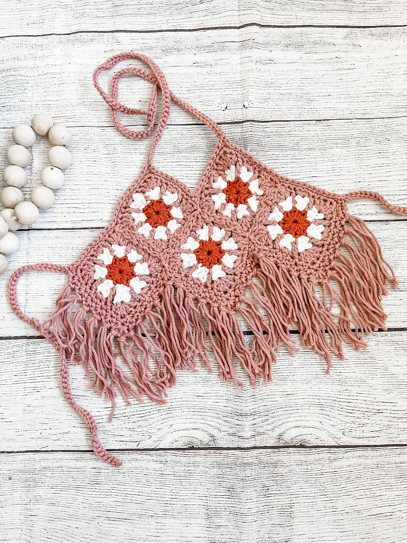 May include: A pink crochet top with white and orange flowers and fringe trim.