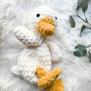 May include: A handmade white crochet duck plush toy with a fluffy texture. The duck has a yellow beak and feet, and black eyes. The toy is lying on a white, fluffy surface.