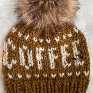 May include: A brown knit hat with the word "COFFEE" in white block letters. The hat features a large, fluffy, brown pom-pom on top. A small, brown tag is sewn on the brim.
