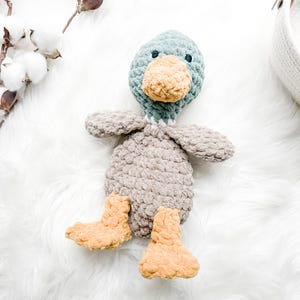 May include: A crocheted plush duck toy with a gray body, teal head, and yellow feet. The duck is sitting on a white fluffy surface.