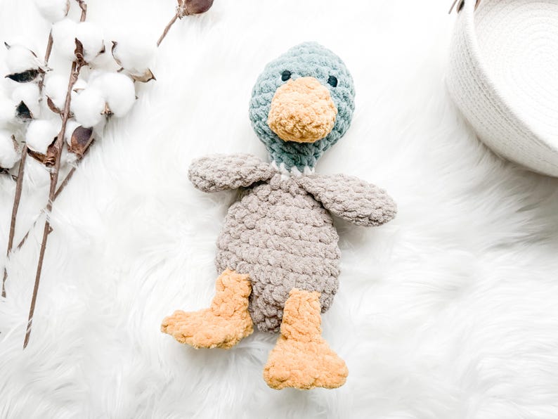 May include: A crocheted plush duck toy with a gray body, teal head, and yellow feet. The duck is sitting on a white fluffy surface.