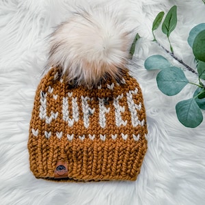 May include: Mustard yellow knit beanie with the word "COFFEE" in white block letters. The hat features a large, fluffy, cream-colored pom-pom on top. A small leather tag is sewn on the brim. The hat is on a white, fluffy surface.