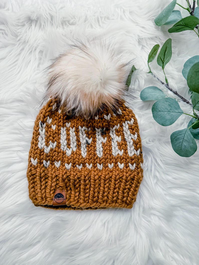 May include: Mustard yellow knit beanie with the word "COFFEE" in white block letters. The hat features a large, fluffy, cream-colored pom-pom on top. A small leather tag is sewn on the brim. The hat is on a white, fluffy surface.
