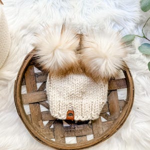 May include: Cream-colored knit hat with two large, fluffy, light brown pom-poms. The hat is displayed on a round, woven wooden tray, set against a backdrop of white faux fur. A small leather tag is sewn on the hat.