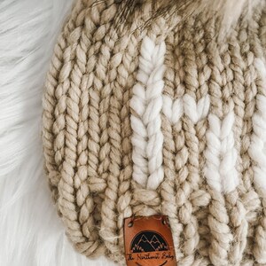 May include: A beige knitted beanie with a white knit pattern and a brown leather patch that says "The Northwest Baby" with a mountain range design.