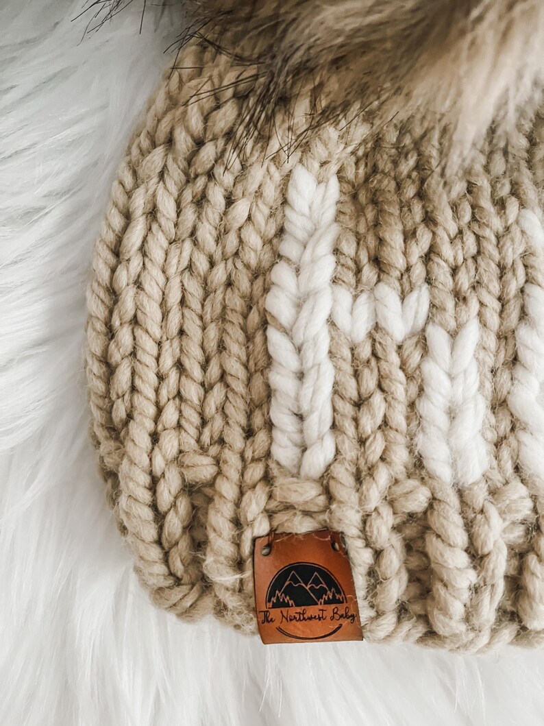 May include: A beige knitted beanie with a white knit pattern and a brown leather patch that says "The Northwest Baby" with a mountain range design.