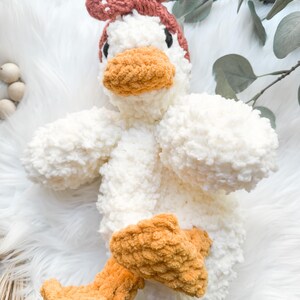 May include: A white crocheted plush duck toy with a brown crocheted bow on its head and orange crocheted feet.