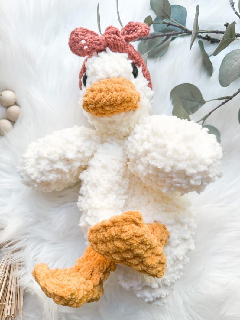 May include: A white crocheted plush duck toy with a brown crocheted bow on its head and orange crocheted feet.