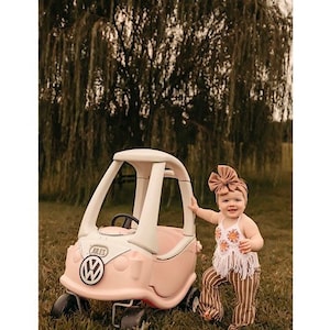 May include: A young child wearing a brown headband and a white crochet top with fringe sits in a pink and white toy car. The car has a white steering wheel and a black VW logo on the front.