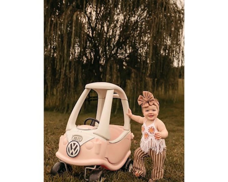 May include: A young child wearing a brown headband and a white crochet top with fringe sits in a pink and white toy car. The car has a white steering wheel and a black VW logo on the front.