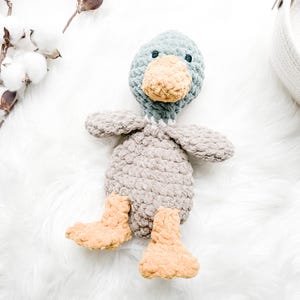 May include: A handmade crochet duck toy in shades of gray, blue, and tan. The duck has a gray body, blue head, tan beak and feet, and small wings. The soft toy is placed on a white, fluffy surface.
