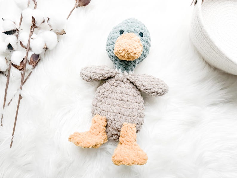 May include: A handmade crochet duck toy in shades of gray, blue, and tan. The duck has a gray body, blue head, tan beak and feet, and small wings. The soft toy is placed on a white, fluffy surface.