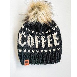 May include: Black knit beanie with the word "COFFEE" in white block letters. The hat features a faux fur pom-pom and a small brown leather tag. The design includes white heart-shaped accents.
