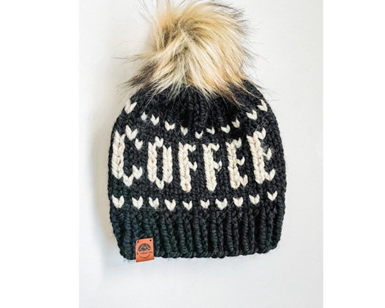 May include: Black knit beanie with the word "COFFEE" in white block letters. The hat features a faux fur pom-pom and a small brown leather tag. The design includes white heart-shaped accents.