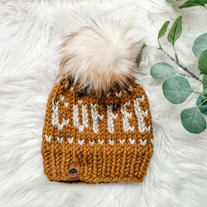 May include: Mustard yellow knit beanie with the word "COFFEE" in white letters. The hat features a fluffy, light brown pom-pom on top and a small leather tag. The beanie is displayed on a white, fluffy surface.