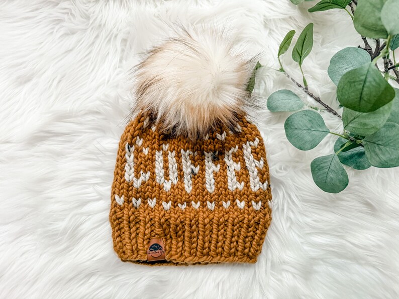 May include: Mustard yellow knit beanie with the word "COFFEE" in white letters. The hat features a fluffy, light brown pom-pom on top and a small leather tag. The beanie is displayed on a white, fluffy surface.