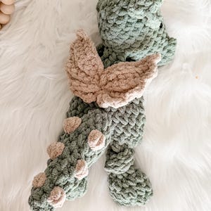 Peut inclure: Un jouet dragon en crochet fait &agrave; la main, dans des tons de vert et de beige. Le dragon pr&eacute;sente un corps textur&eacute;, des ailes beiges et de petits accents beiges le long de sa queue. Le jouet est pos&eacute; sur une surface blanche et duveteuse.