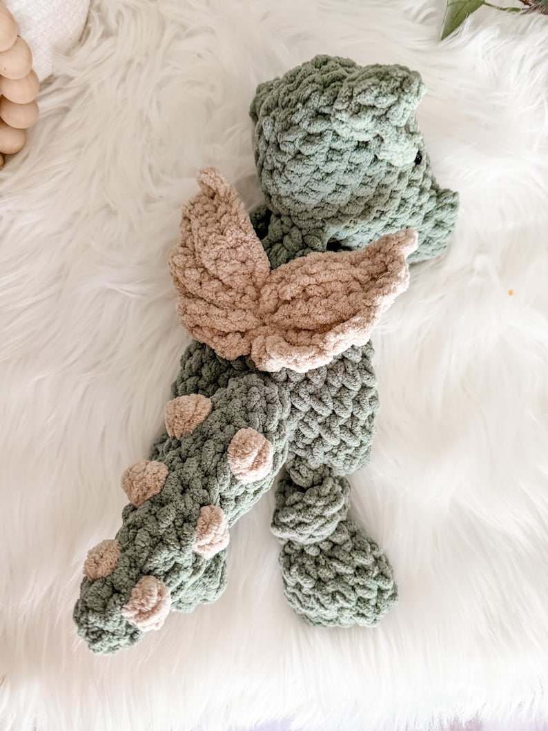 Peut inclure: Un jouet dragon en crochet fait &agrave; la main, dans des tons de vert et de beige. Le dragon pr&eacute;sente un corps textur&eacute;, des ailes beiges et de petits accents beiges le long de sa queue. Le jouet est pos&eacute; sur une surface blanche et duveteuse.