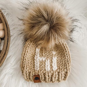 May include: A beige knitted hat with a white "Hi" embroidered on the front. The hat has a large brown faux fur pom pom on top.
