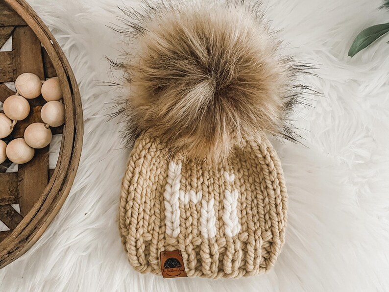 May include: A beige knitted hat with a white "Hi" embroidered on the front. The hat has a large brown faux fur pom pom on top.