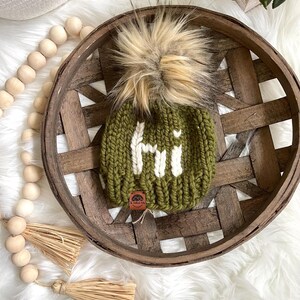 May include: A green knitted beanie with a white "Hi" embroidered on the front. The beanie has a fluffy brown and white pom pom on top.