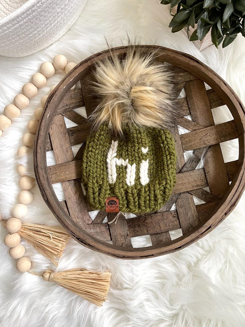 May include: A green knitted beanie with a white "Hi" embroidered on the front. The beanie has a fluffy brown and white pom pom on top.