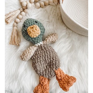May include: A crocheted stuffed duck toy with a green head, brown body, and orange feet. The duck is lying on a white fluffy surface.