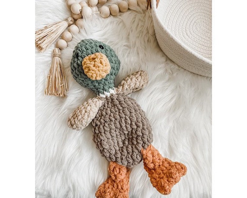 May include: A crocheted stuffed duck toy with a green head, brown body, and orange feet. The duck is lying on a white fluffy surface.