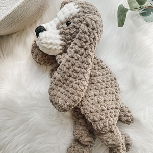 May include: A crocheted plush dog toy in brown and white. The dog is lying on a white fluffy surface.