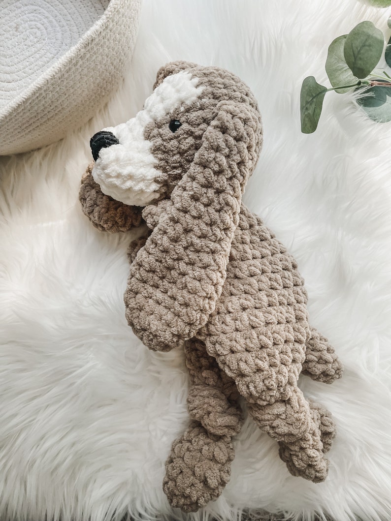 May include: A crocheted plush dog toy in brown and white. The dog is lying on a white fluffy surface.